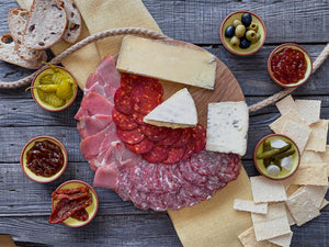 Three types of sliced cured meats and three cheeses on a circular wooden board. Six clay dishes, two containing pickles, 2 of chutney and one each of olives and sundried tomatoes. Four slices of sourdough bread and an array of crackers.