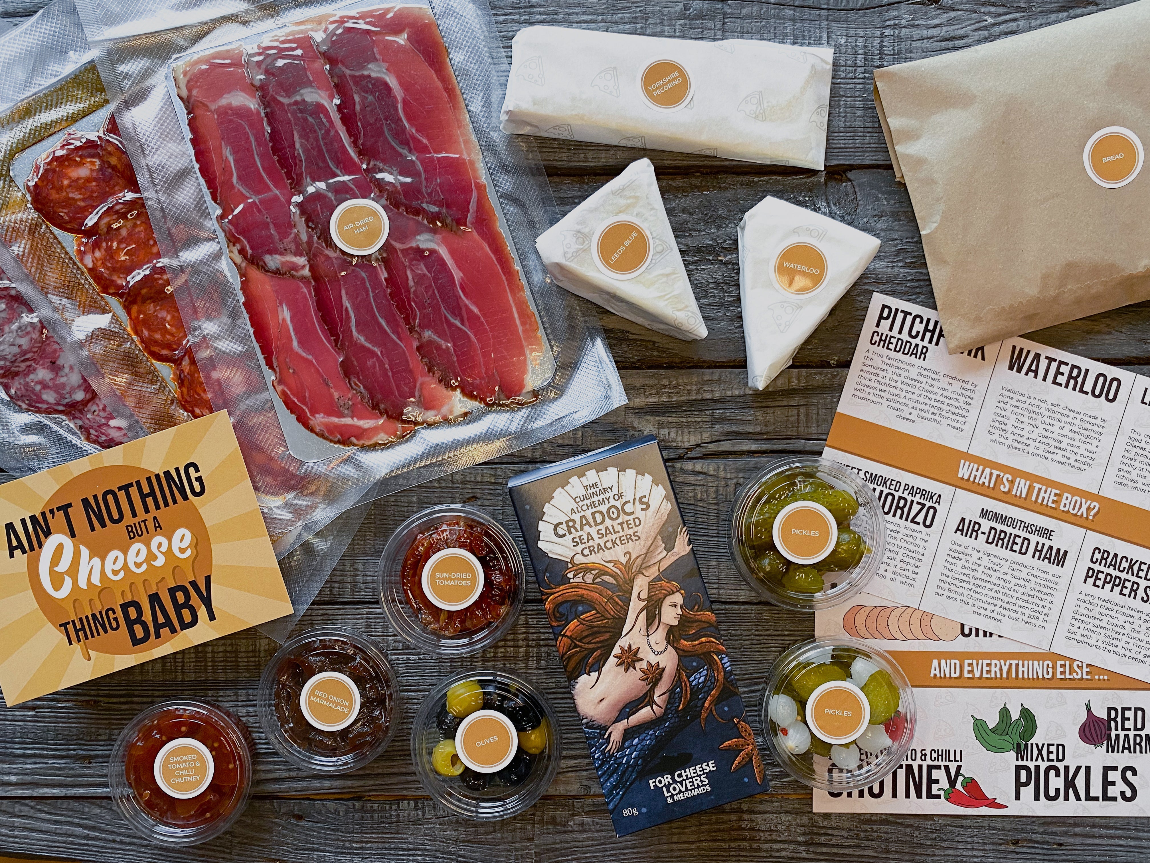 Three types of sliced cured meats vacuum sealed individually and three individually wrapped portions of cheese. A packet of crackers, and individual portion pots of olives, sun dried tomatoes, red onion marmalade, smoked tomato and chilli chutney, and two variations of pickles, all on a wooden background. There's an A5 descriptive 'What's in the box' card visible, which describes the produce in more detail and an A6 graphic postcard with the words 'Ain't nothing but a cheese thing baby'.