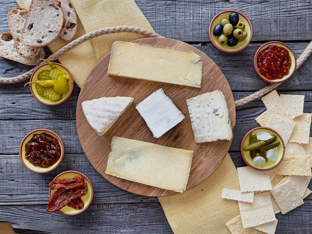 5 cheeses on a circular wooden board. Six clay dishes, two containing pickles, 2 of chutney and one each of olives and sundried tomatoes. Four slices of sourdough bread and an array of crackers.