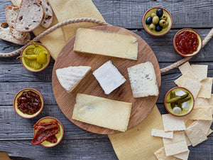 5 cheeses on a circular wooden board. Six clay dishes, two containing pickles, 2 of chutney and one each of olives and sundried tomatoes. Four slices of sourdough bread and an array of crackers.