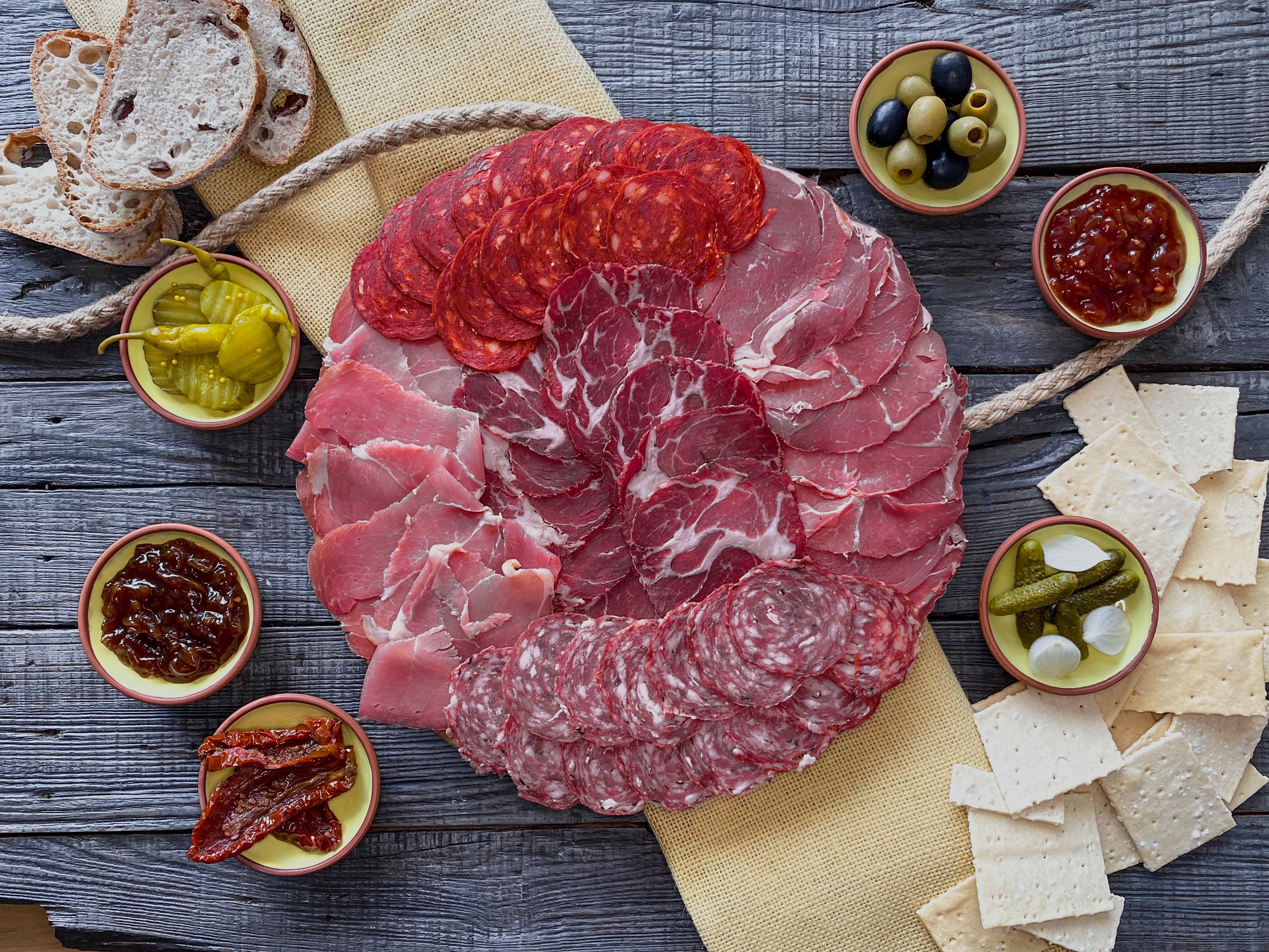 Five types of sliced cured meats on a circular wooden board. Six clay dishes, two containing pickles, 2 of chutney and one each of olives and sundried tomatoes. Four slices of sourdough bread and an array of crackers.